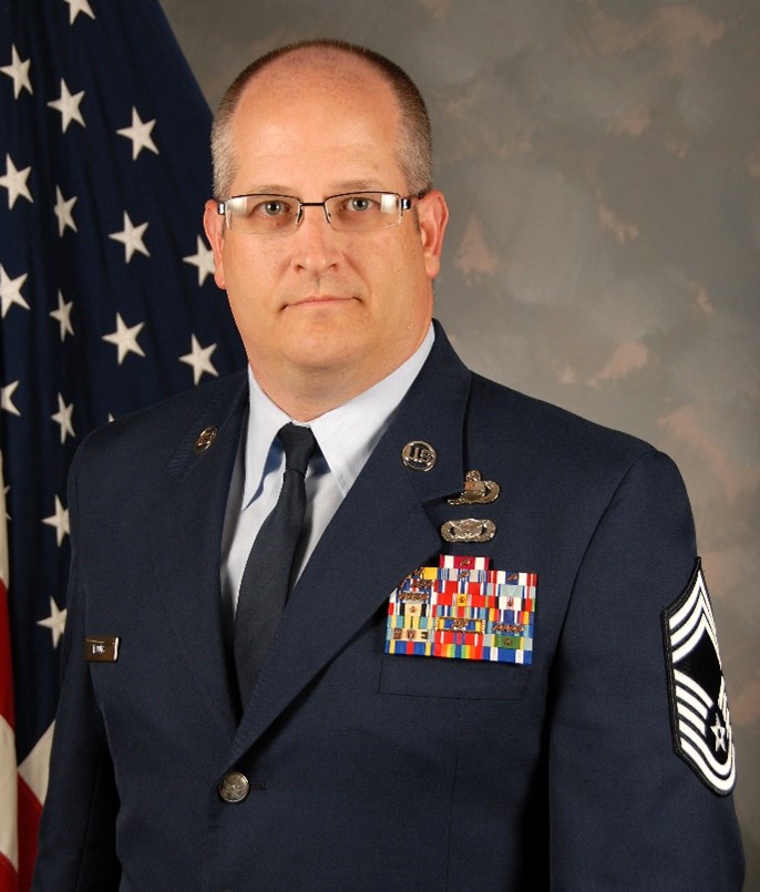 A man wearing glasses in Air Force dress Blues poses for a portrait in front of the American Flag. 